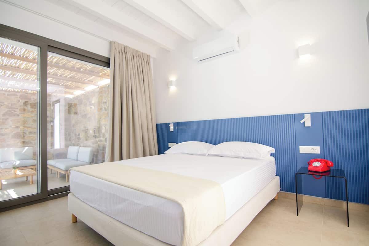 Habitación de hotel con cama, puerta de vidrio que da a un balcón, teléfono rojo y aire acondicionado de pared.