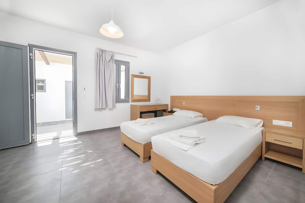 Una habitación de hotel moderna con dos camas individuales, una mesita de noche de madera, un escritorio con una silla, un espejo grande y un cerramiento de vidrio corredera que da a un balcón.
