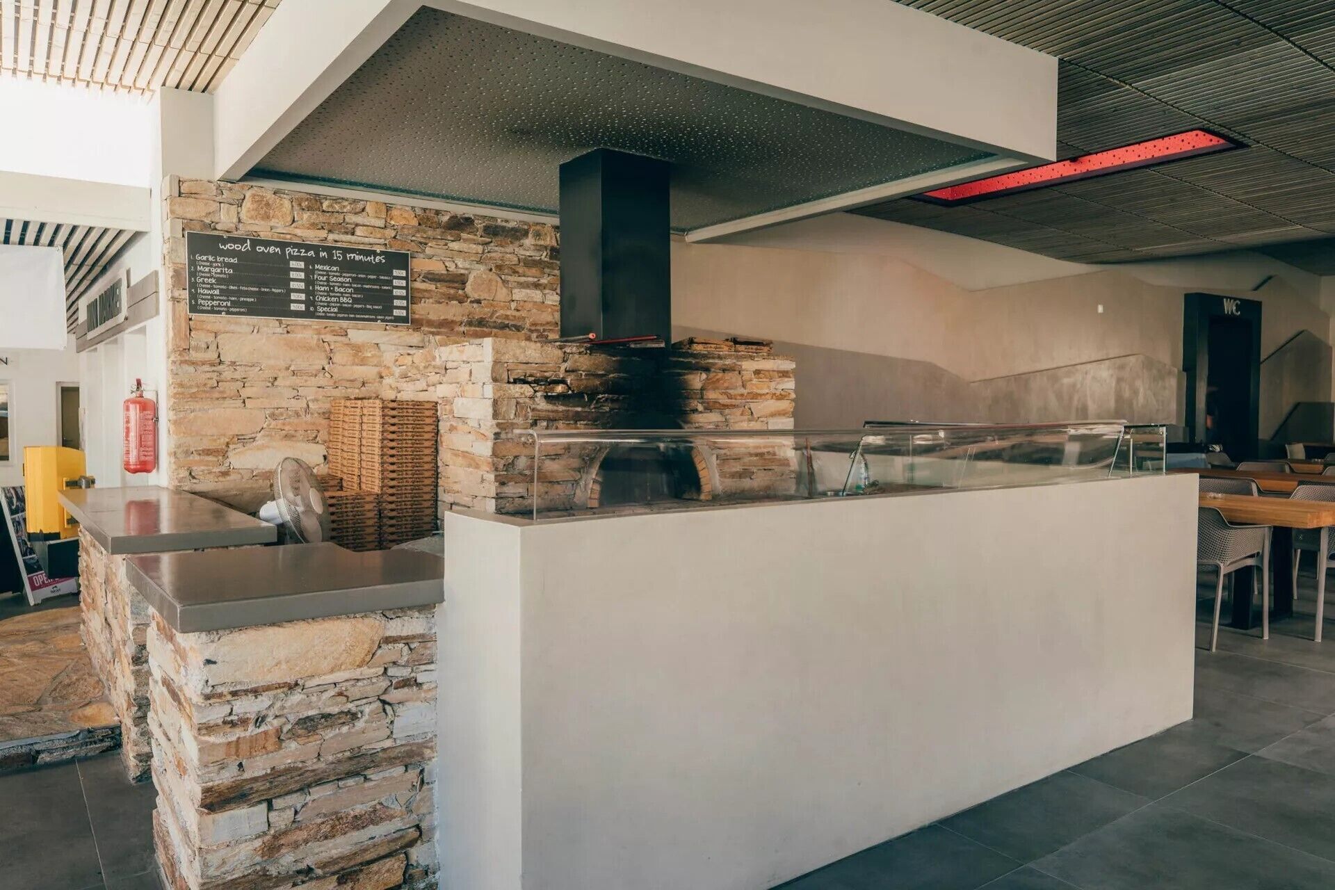 A modern kitchen area with a stone wall, a wood oven, and a menu board listing pizza options.