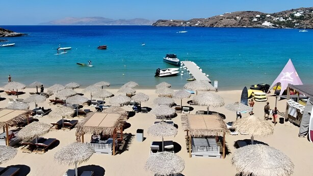 A beach with thatched umbrellas and sun loungers, a pier extending into the sea, and boats anchored nearby. - FarOut BeachClub - Rooms & Camping (Ios)
