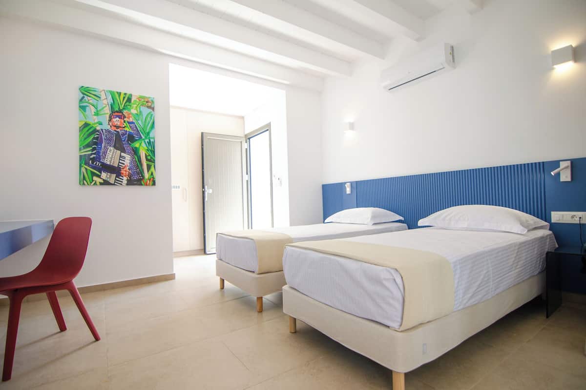 Habitación de hotel con dos camas, una silla roja, un cuadro en la pared y una puerta que da a un balcón.
