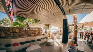 A modern outdoor lounge area with a stone wall, seating with circular and rectangular cushions, and a canopy overhead. - FarOut BeachClub - Rooms & Camping (Ios)