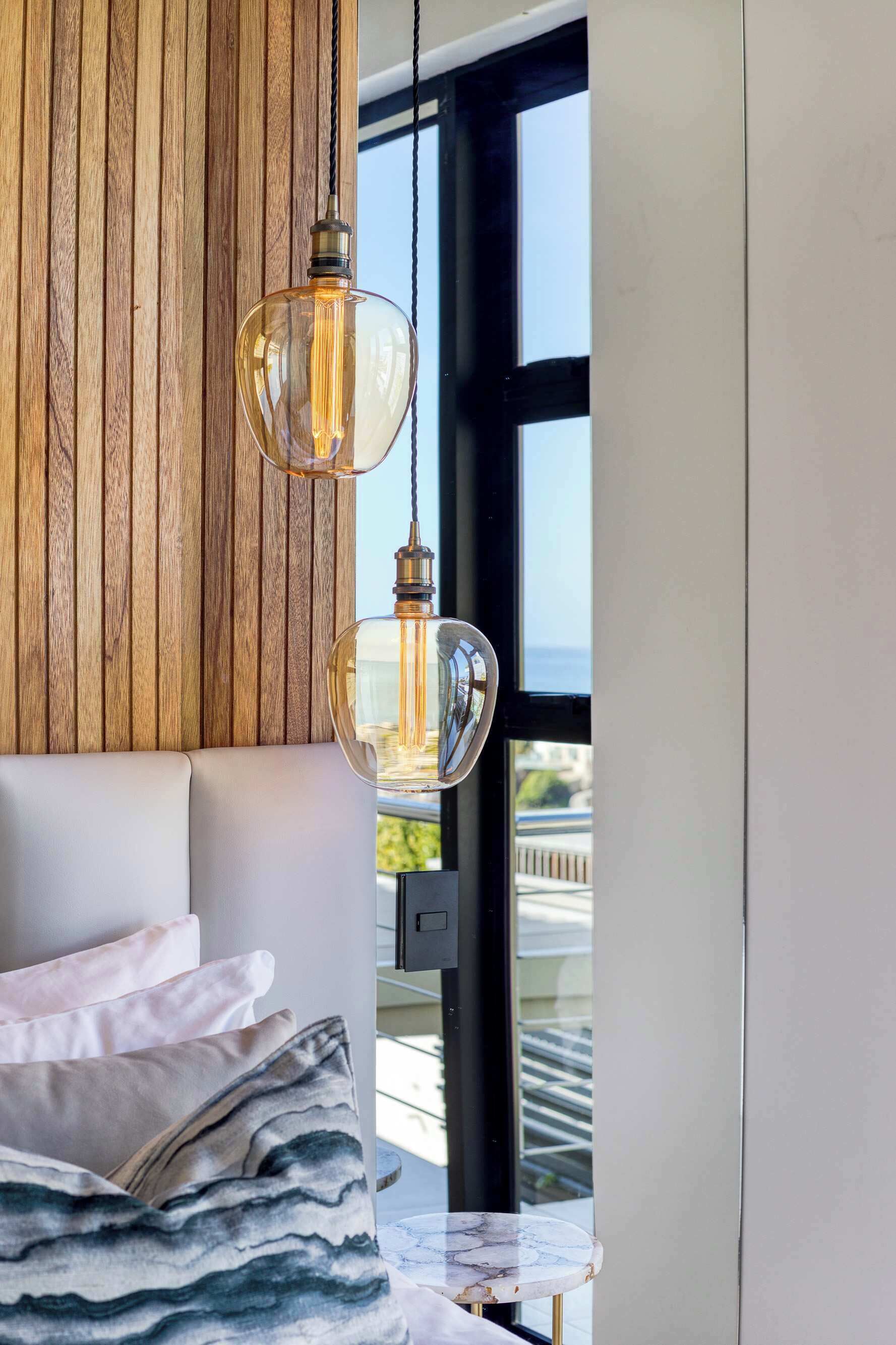 Une chambre moderne avec un mur en bois, une grande fenêtre, un lit avec une literie blanche et grise, et deux lampes suspendues.