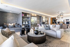 A modern living room with a grey sofa, a round coffee table, and a kitchen area in the background. - Barley Beach Luxury Penthouse - Camps Bay (Cape Town)