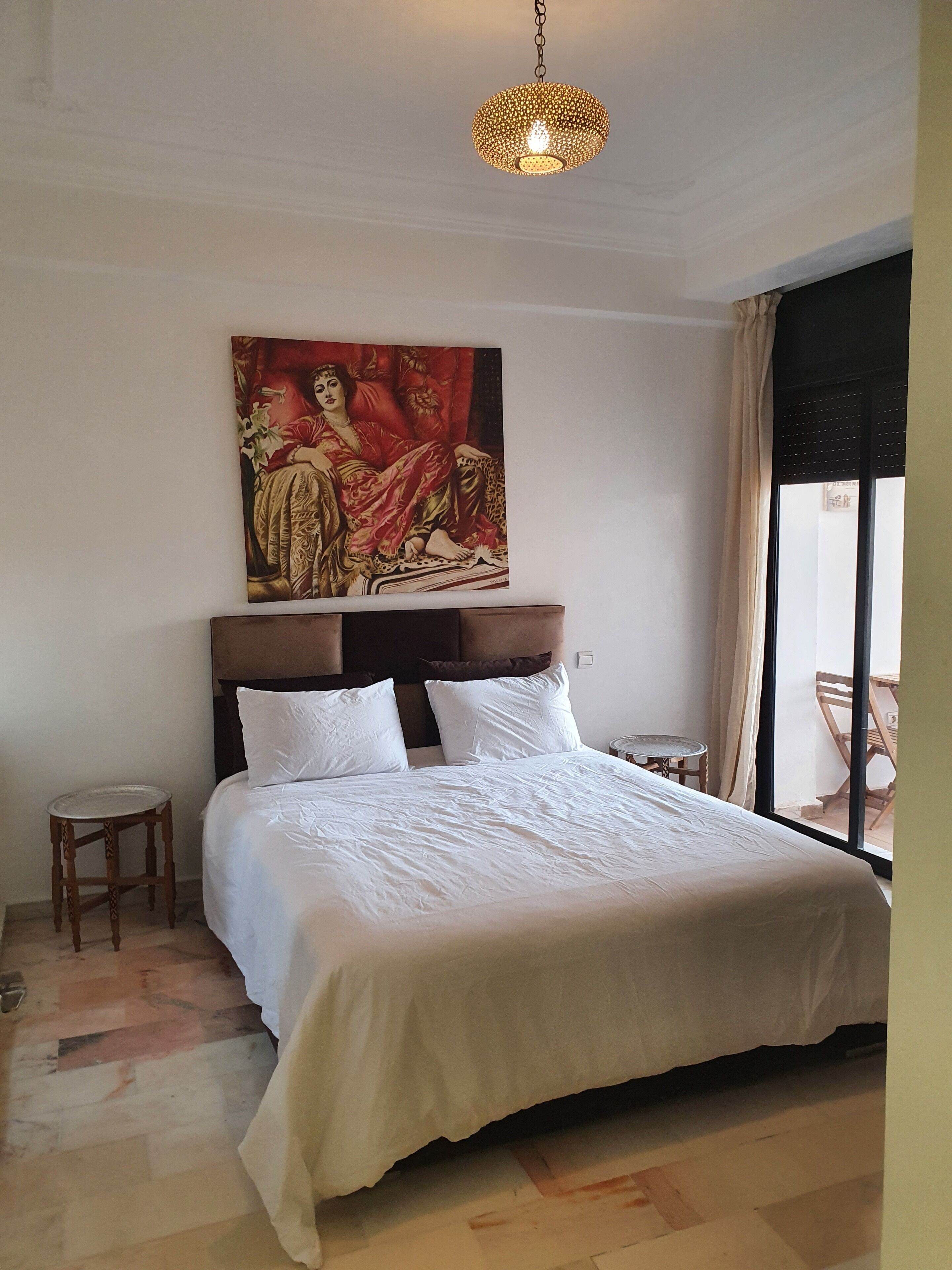 A bedroom with a large bed, a headboard with a painting, a small table, and a chair.
