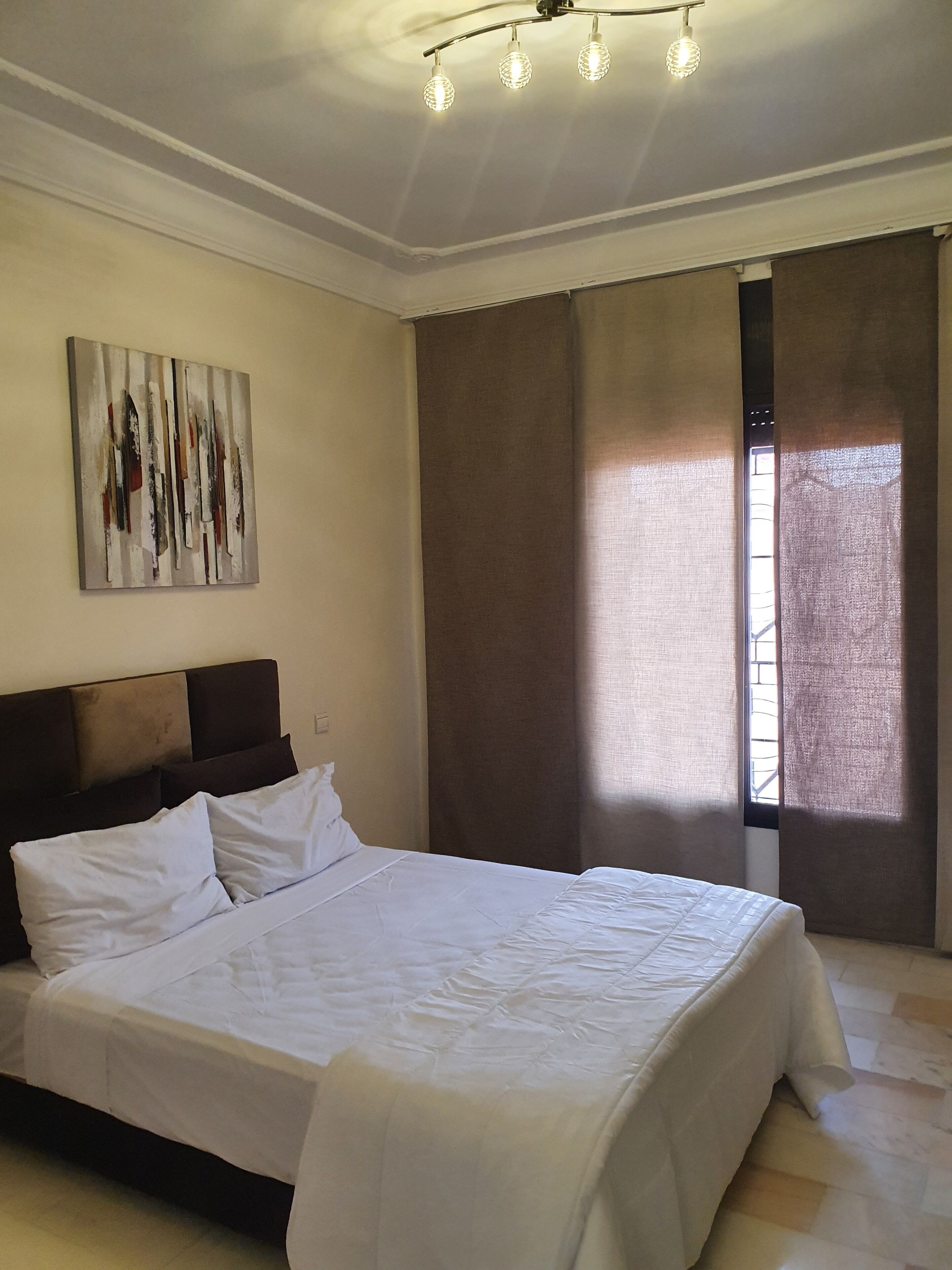 A bedroom with a large bed, a ceiling light fixture, a window with curtains, and a wall-mounted artwork.
