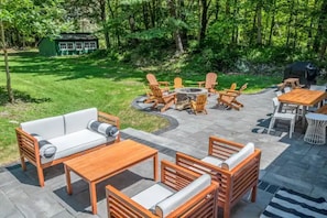 Eine Außenpatio-Terrasse mit Gartenmöbeln aus Holz, darunter eine Couch, Stühle und Tische, umgeben von einem üppigen Grünbereich.