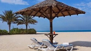 A beach umbrella with a table and two lounge chairs on a sandy beach with palm trees and the ocean in the background.