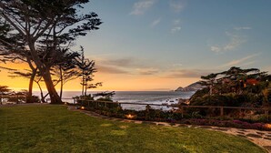 A coastal landscape with a grassy area, trees, and a view of the ocean at sunset.