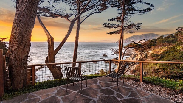 A patio with stone flooring, two chairs, and a view of the ocean and coastal landscape.