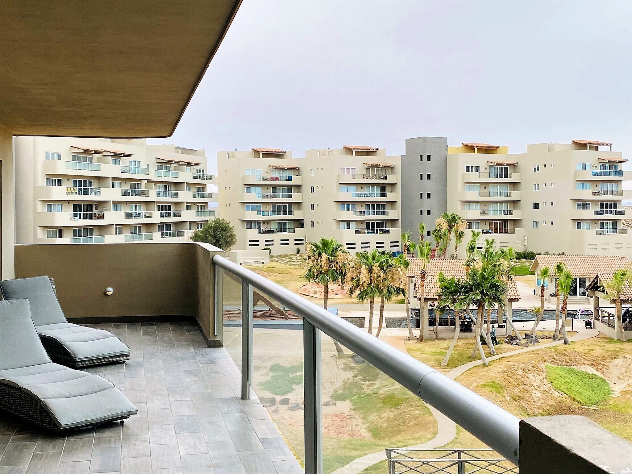 Ein Balkon mit Liegestühlen, Blick auf ein Wohngebiet mit mehreren Mehrfamilienhäusern und ein kleiner Garten mit Palmen.