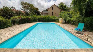 Una piscina rectangular con borde de piedra, rodeada por un jardín y una casa al fondo.