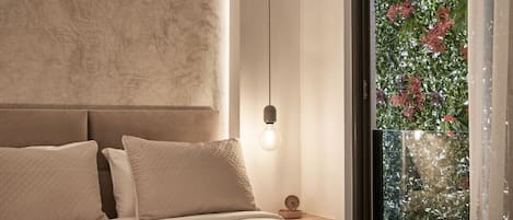 A bedroom with a bed, a nightstand, a wall clock, and a window with a view of greenery.