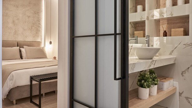 A modern bedroom with a large bed, bedside tables, and a bedside lamp. The bathroom features a large sink, mirror, and shelves with toiletries.