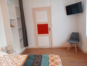 A room with a bed, a chair, a wardrobe, a door with a red panel, and a flat-screen TV mounted on the wall. - Quiet & Bright Apartment (Le Creusot)