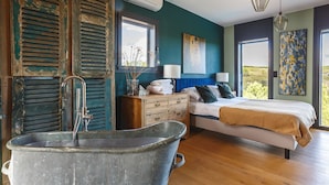 A bedroom with a large tub, a bed with blue curtains, and a view of greenery outside.