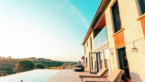 A modern villa with a wooden deck, a pool, and lounge chairs.