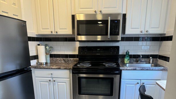 A modern kitchen with white cabinets, a stainless steel refrigerator, a microwave, and an oven. - Bella Is A Happy Place. (Lehigh Acres)