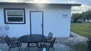 A small building with a white exterior, a door, and a window. There is a table with chairs outside, and a sign on the wall. - Bella Is A Happy Place. (Lehigh Acres)