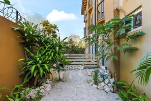 Stroll through the charming entrance, lined with lush greenery and a wooden fence, to the building's inviting facade with multiple windows and balconies.