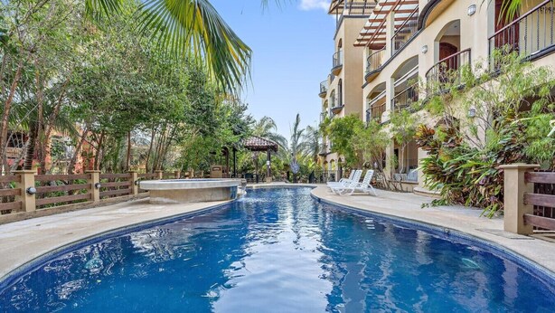 Uma piscina de borda curva, rodeada por vegetação exuberante e um edifício com várias andares e varandas.