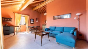 Un salon avec un canapé bleu, des poutres apparentes en bois au plafond, un coin repas avec des chaises et une table, et un coin cuisine avec une étagère et un meuble.