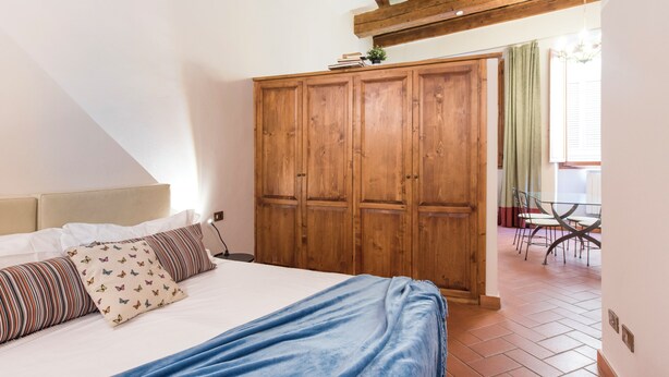A bedroom with a bed, wooden wardrobe, and a dining area with chairs and a table.