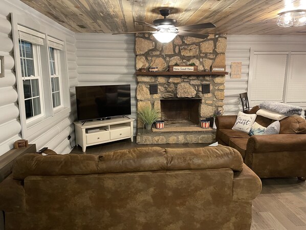A log cabin interior with a stone fireplace, a flat-screen TV on a white cabinet, and two brown sofas. - Real Log Cabin, new remodel and Great Location (Pigeon Forge)