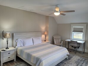 Une chambre à coucher comprenant un lit, des tables de chevet, un bureau avec une chaise, un ventilateur de plafond et une fenêtre avec des stores.