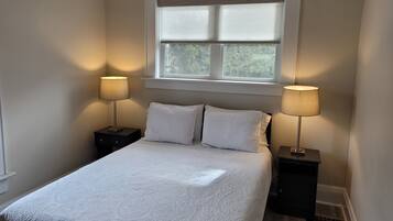 A bedroom with a bed, two bedside tables, a window with blinds, and a nightstand with a lamp.