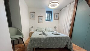 Un dormitori amb un llit, una cadira, una finestra i reproduccions botàniques emmarcades a la paret.