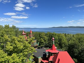 A view of a castle-like building with multiple towers, surrounded by dense green trees and a large body of water in the background. - Auberge Château Bahia (Escuminac)