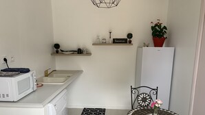Een compacte keuken met een magnetron, spoelbak en een klein eettafeltje met een decoratief tegelblad. Boven de spoelbak zijn planken met decoratieve items en een potplant.