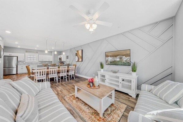 Una cocina moderna con muebles blancos, un comedor con mesa y sillas de madera, y una sala de estar con sofás a rayas y una mesa de centro.