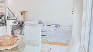 En moderne stue med et glasbord, hvide sofaer og en trappe, der fører til en etage ovenpå.