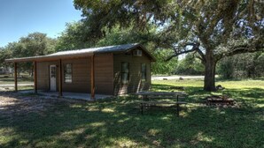 A rustic cabin with a covered porch, a bench, and a picnic table in a wooded area. - Zubers - CA -F Cabin F (Concan)