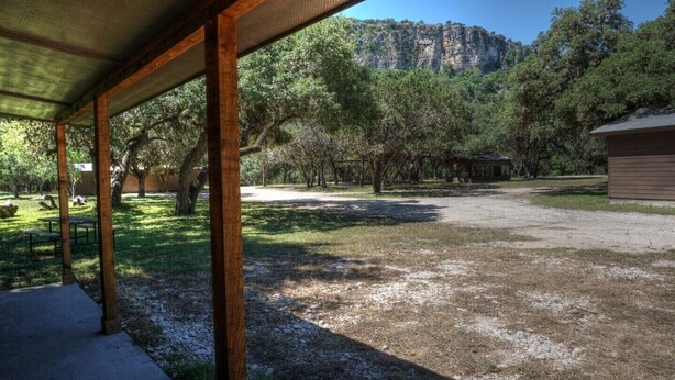 A covered outdoor area with a gravel path, picnic tables, and a small building in the background. - Zubers - CA -F Cabin F (Concan)