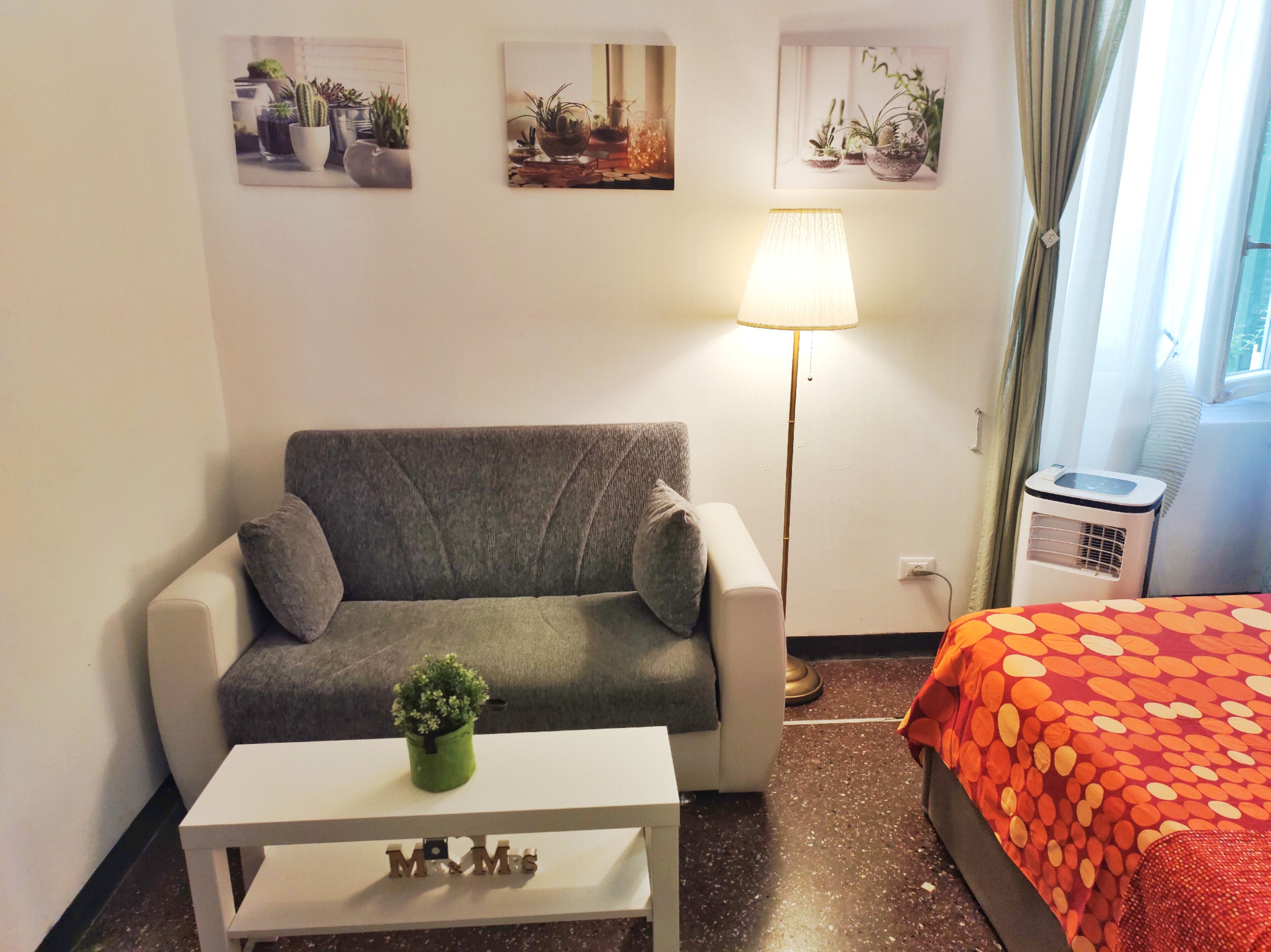 Una habitación con un sofá gris, una mesita blanca con una planta en maceta, una lámpara de pie, una cama con una manta de lunares y tres cuadros en la pared.