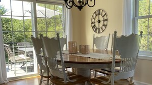 Une salle à manger avec une table en bois, des chaises blanches, un lustre et une grande fenêtre donnant sur un paysage verdoyant.