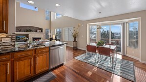 A spacious kitchen with wooden cabinets, a granite countertop, and a built-in dishwasher. There is a dining area with a table and chairs, and large windows providing natural light. - Aurora Manor with Breathtaking Views (Aurora)