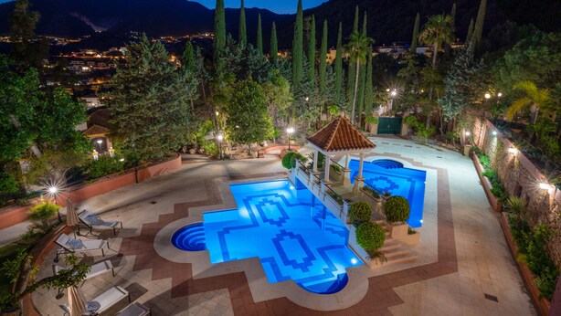 Uma área de piscina no terraço com design de azulejos geométricos, cercada por espreguiçadeiras e guarda-sóis, com montanhas ao fundo.