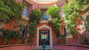 Um pátio com uma fonte, rodeado por vegetação exuberante e fachada rosa de um edifício.