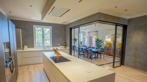 Uma cozinha moderna com uma ilha central, área de jantar com cadeiras azuis e vista para uma sala de estar através de uma porta de vidro.
