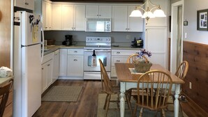 Una cocina con armarios blancos, estufa, refrigerador, sillas de madera y una araña.