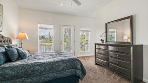 Ein Schlafzimmer mit einem großen Bett, einem Schminktisch und einem Spiegel. Zwei Fenster mit Jalousien sind vorhanden, und durch eines der Fenster ist ein Balkon zu sehen.