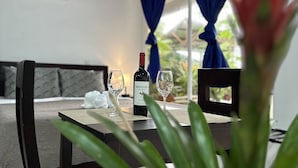 A dining area with a table set for two, a bottle of wine, and two glasses. There is a plant in the foreground, and the room has a modern design with a window and curtains. - Hotel Serendipity tortuguero (Colorado)