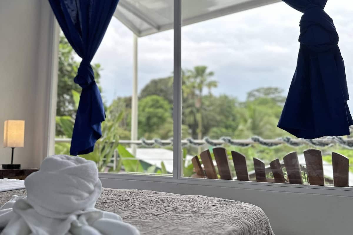Ein Hotelzimmer mit einem Bett, einem Fenster mit blauen Vorhängen und Blick auf Grünfläche.