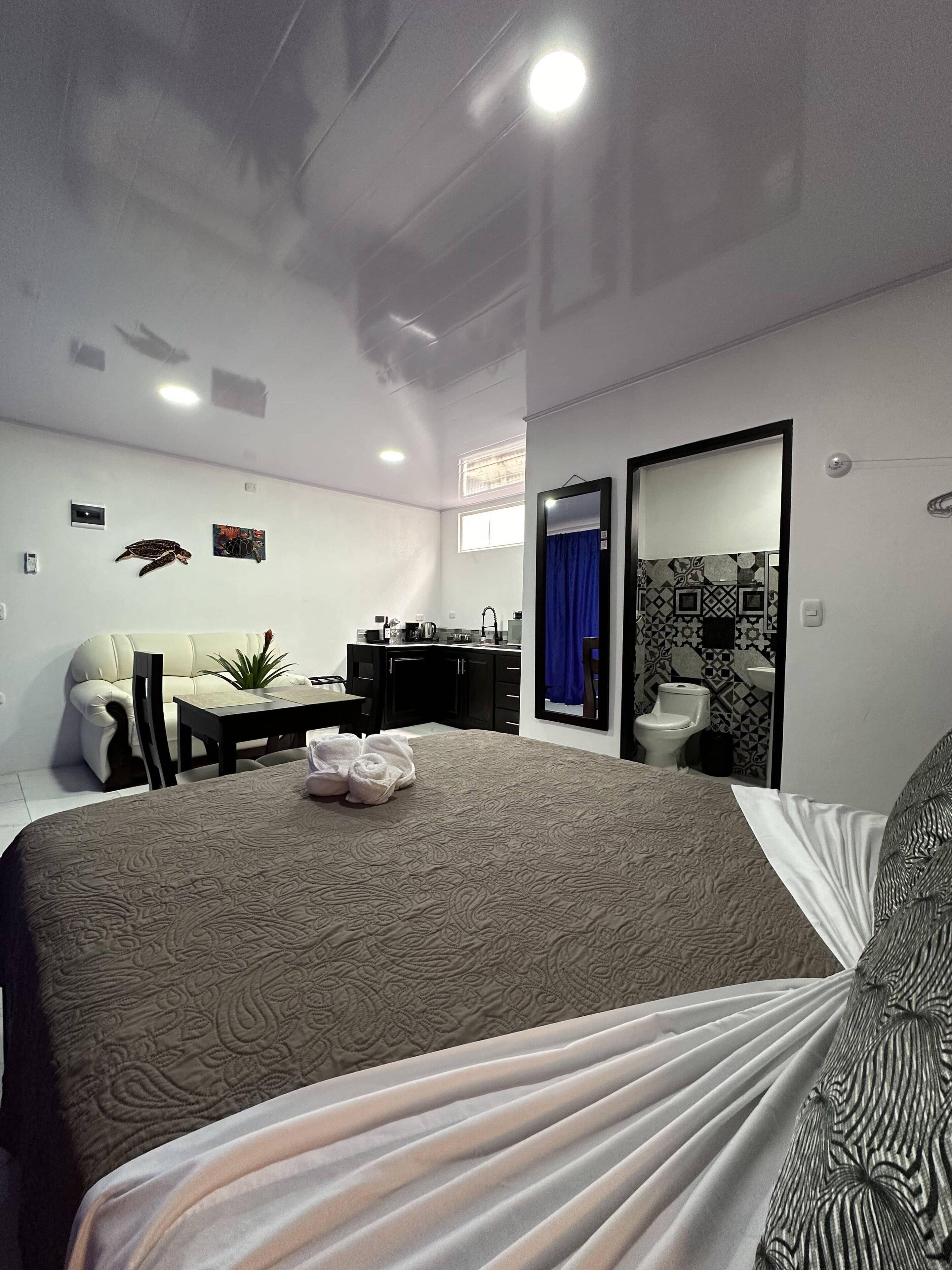 Una habitación de hotel moderna con una cama grande, un escritorio, una silla, una mesita, una planta en maceta y un baño con espejo y lavabo.