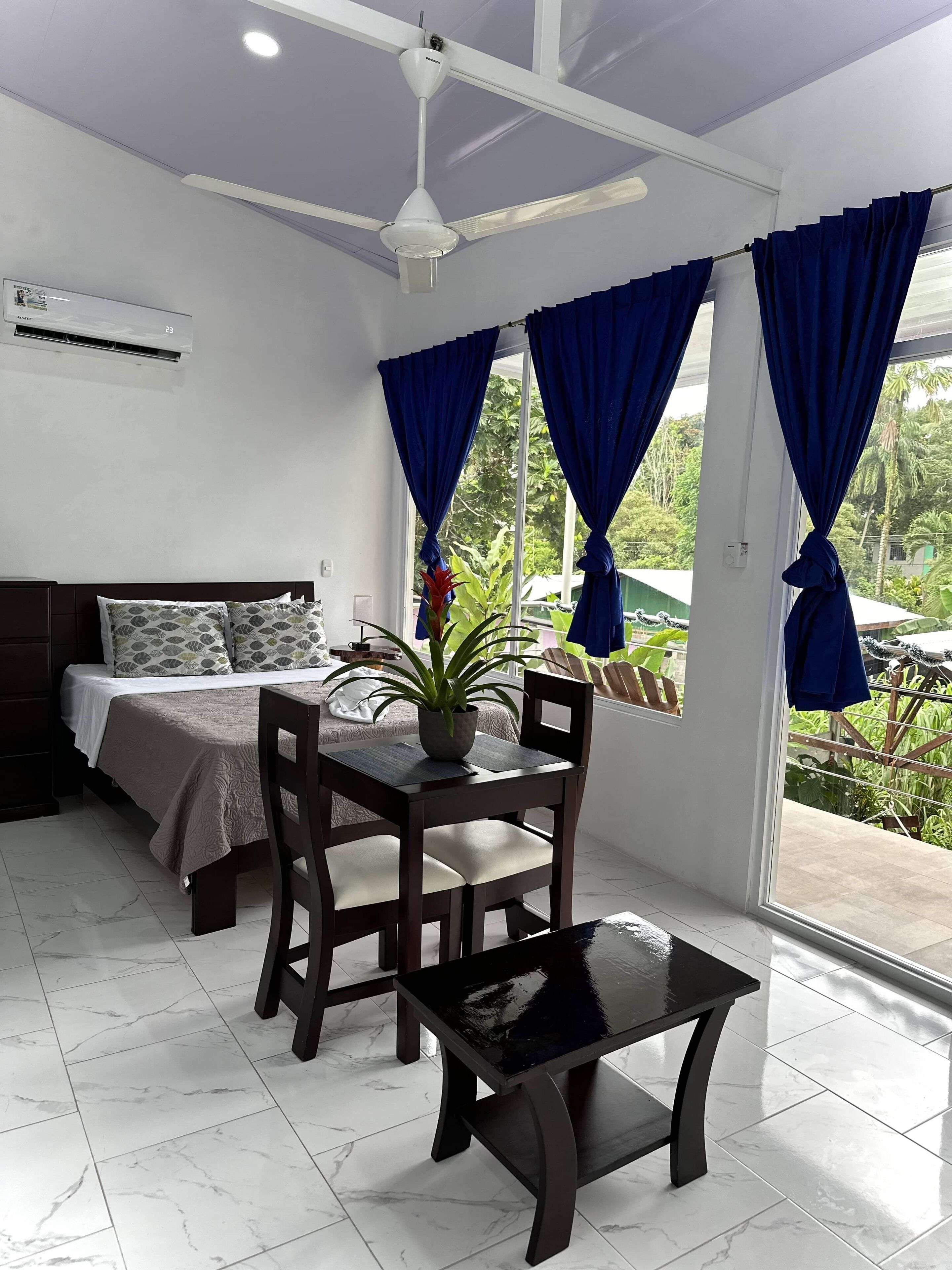 A hotel room with a bed, a dining table with chairs, a ceiling fan, and a window with curtains.