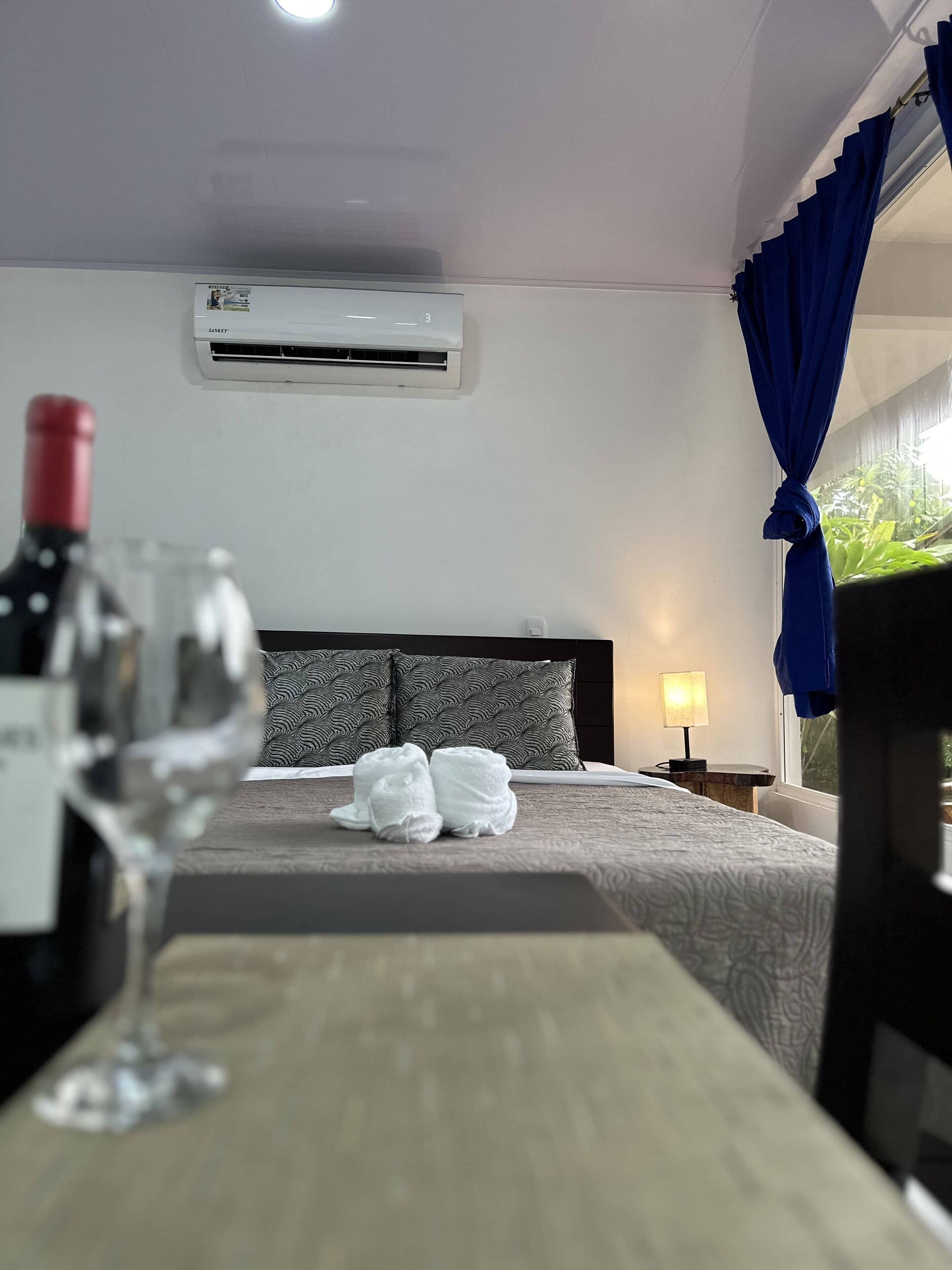 A hotel room with a bed, a nightstand, a lamp, a window with blue curtains, and an air conditioning unit.